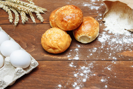 Sesame buns on a brown wooden table with a paper bag of flour, white eggs and a bunch of wheat, the...