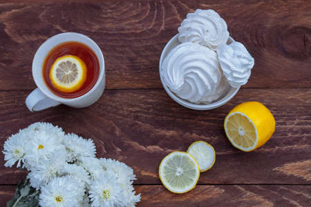 White large meringues in a bowl with a Cup of tea with lemon slices and a bouquet of white chrysanthemums on a brown wooden background top view, flat composition, concept of low-fat sweets, Breakfastの写真素材