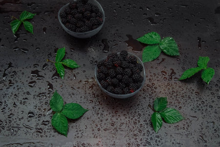 ripe blackberries in glass salad bowls on a black table with water drops and green leaves top view, berry harvest concept, vitamins, ingredientsの写真素材