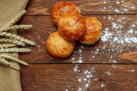 Round sesame buns on a brown wooden background with a linen cloth, a bunch of wheat and spilled...