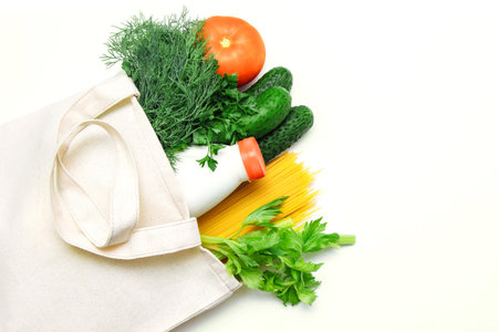 White cloth bag with vegetables, tomatoes, cucumbers, dill, parsley, celery, milk and spaghetti on a...