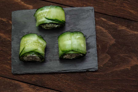 Green vegetable rolls with cucumber cheese on a black silicon-graphite saucer top view on a brown wooden background, with an empty space for text concept of holiday snacks, buffet, vegetarian foodの写真素材