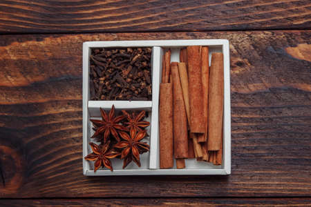 Anise stars, cinnamon sticks and cloves in white cardboard box on brown wooden background closeup view from above, concept of spices for baking and drinks, mulled wine, Christmas and healthy foodの写真素材