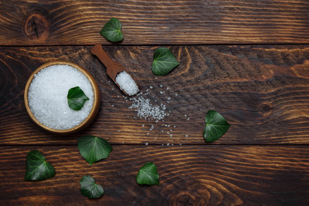 White sea salt in a bowl with a spoon and green ivy leaves, top view flat composition with empty space for text on brown wooden background, spa treatment concept, bath and sauna, natural scrubの写真素材