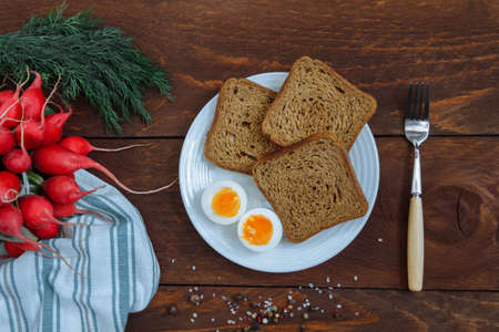 Slices of rye bread on white plate with an egg and bunch of radish and dill on brown wooden background with fork and scattered salt and pepper spices, concept of cooking healthy food, breakfastの写真素材