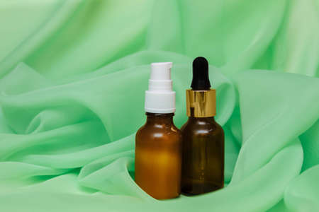 Two cosmetic glass bottles with pipette and dispenser on the background of green silk fabric with pleats, copy space, anti-aging and acne skin care concept, product advertising templateの写真素材