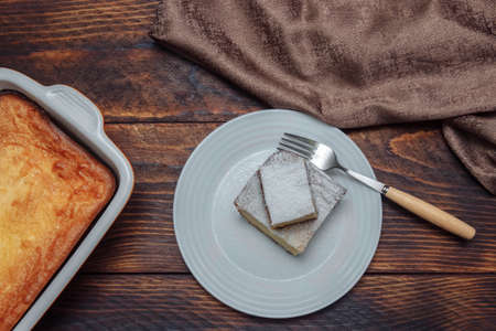 Cottage cheese casserole in baking dish and a slice in a white plate with powdered sugar and fork on brown wooden background with napkin, top view, healthy breakfast concept, dessert cooking recipeの写真素材