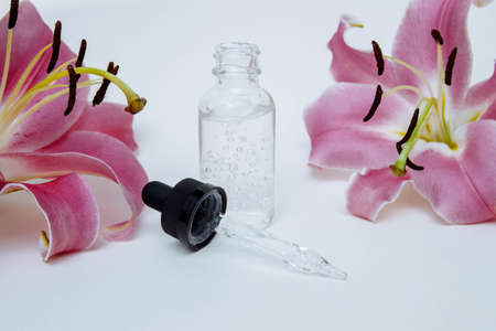 Cosmetic glass bottle with hyaluronic acid and pipette on white background with pink lily flowers close-up, skin care concept, organic cosmetics, moisturizing and rejuvenation of face, copy spaceの写真素材