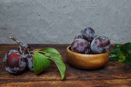 Ripe, fresh plums in a wooden bowl and on a branch with green leaves close-up on a brown kitchen table and gray plastered walls, the concept of organic harvest, rustic, vitamins and vegan ingredientsの写真素材