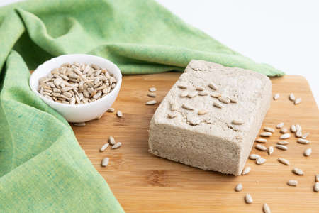 A piece of sunflower halva and sunflower seeds on a wooden cutting board next to a green towelの写真素材