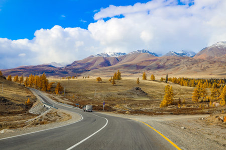 A winding road, snow-capped mountain peaks, the blue sky of the autumn Altai, a picturesque region, nature, rivers and roads in autumn. Yellow foliage, clouds. Travel, tourism in Russia, auto tourism, tours.の写真素材