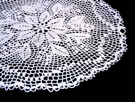 A part of isolated crocheted white doily with a pattern of flowers on a black background. Round decorative cotton doily. Side viewの写真素材