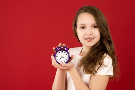 beautiful teenage girl smiling with an alarm clock in hands on a red background. teenager looks at the camera, right side viewの写真素材
