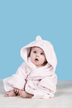 cute baby sitting on a blanket in pink bathrobe with hood and with surprise looks aside. child about six month with big blue eyes on blue, isolated. family love and careの写真素材