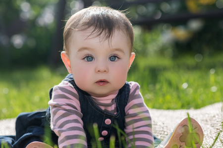 cute little baby girl with big blue eyes and long eyelashes sitting on a green grass on the nature in the park, outdoorの写真素材