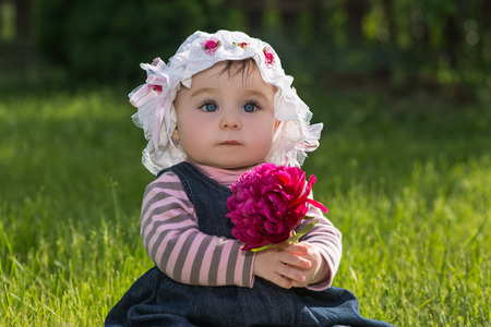 cute little baby girl with big blue eyes in a summer hat with flower in her hands. child sitting on a green grass on the nature in the park, outdoorの写真素材