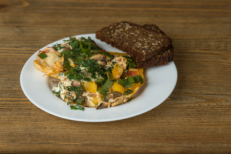 scrambled eggs with ham, mushrooms, tomatoes and slices of bread for breakfast on a plate on a wooden tableの写真素材