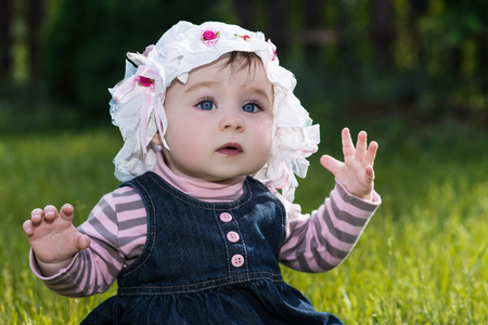 cute little baby girl with big blue eyes in a summer hat sitting on a green grass on the nature in the park, outdoorの写真素材