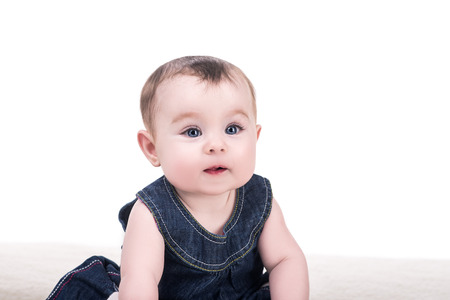 portrait of cute baby girl with big blue eyes and long eyelashes, isolated on white. adorable child girl in blue dress sitting on blanket and smilingの写真素材