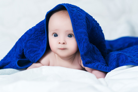 Cute baby girl or boy after shower with towel on head in white sunny bedroom. Child with big blue eyes relaxing in bed after bathの写真素材