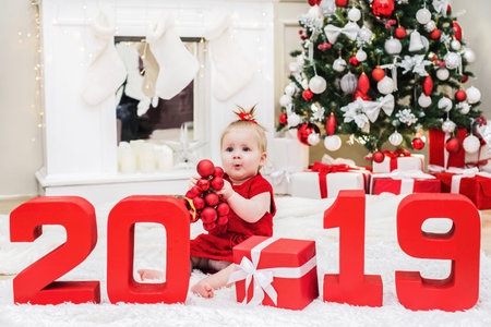 Surprised cute baby girl sits in a red dress with red numbers 2019 in a Christmas and New Year room...