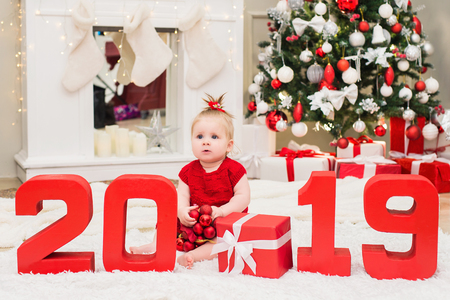 Surprised cute baby girl sits in a red dress with red numbers 2019 in a Christmas and New Year room over Christmas tree, balls and gift boxes backgroundの写真素材