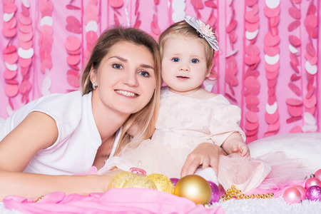 Portrait of happy young mother with her cute baby girl on the soft blanket with Christmas balls and beads, in a festive pink interior. New Year and Christmas holidays.の写真素材