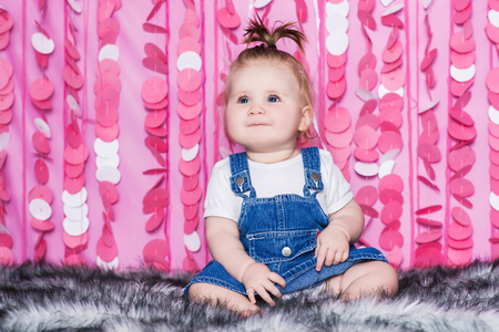 Cute little baby girl with big blue eyes in a blue jeans dress sits on a soft grey blanket, in a festive pink interiorの写真素材