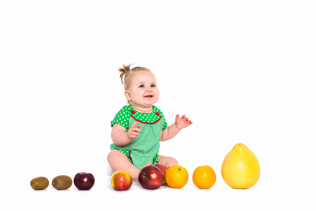 Happy baby girl with big blue eyes in green dress sitting with fruits and vegetables, isolated backgroundの写真素材