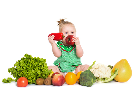 Smiling baby girl sitting surrounded by fruits and vegetables and eating an apple and pepper, isolated over white background. The concept of healthy food and happy lifeの写真素材
