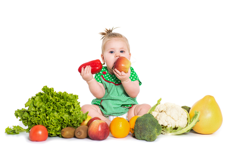 Smiling baby girl sitting surrounded by fruits and vegetables and eating an apple and pepper, isolated over white background. The concept of healthy food and happy lifeの写真素材
