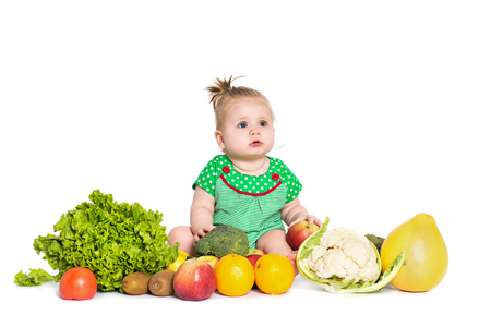Cute baby girl sitting surrounded by fruits and vegetables, isolated over white background. The concept of healthy food and happy lifeの写真素材