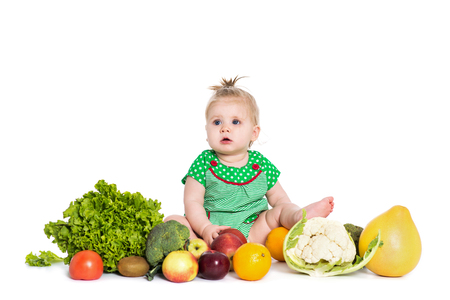 Cute baby girl sitting surrounded by fruits and vegetables, isolated over white background. The concept of healthy food and happy lifeの写真素材
