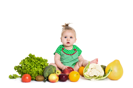 Cute baby girl sitting surrounded by fruits and vegetables, isolated over white background. The concept of healthy food and happy lifeの写真素材