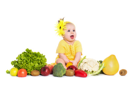 Cute baby girl in yellow dress with flower bow on head sitting surrounded by fruits and vegetables, isolated over white background. Funny smiling child keep an orange in her hands and show a tongue. The concept of healthy food, diet and happy lifeの写真素材