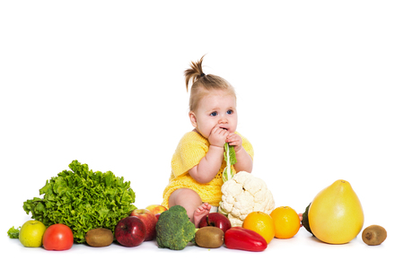 Cute baby girl are sitting surrounded by fruits and vegetables, isolated over white background. Funny child in yellow dress with flower bow on head trying to bite a cabbage. The concept of healthy food, diet and happy lifeの写真素材