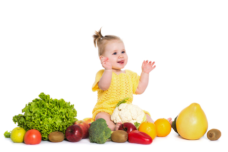 Cute baby girl are sitting surrounded by fruits and vegetables, isolated over white background. Funny smiling child  in yellow dress with flower bow on head show a tongue. The concept of healthy food, diet and happy lifeの写真素材