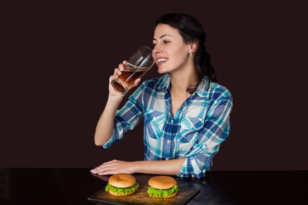 Happy smiling young woman sitting at a wooden table with a mug of beer and hamburgers or sandwiches,...