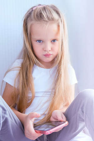 little girl looks at her phone, unhappy sitting at home by the window.Children's dependence on gadgetsの写真素材