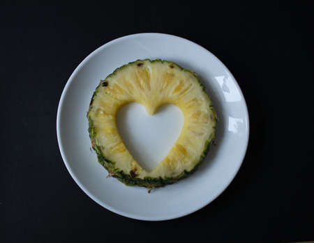 A slice of pineapple with a heart-shaped cut on a white saucer on a black background, close-upの写真素材