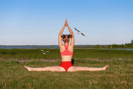 Backwards view of young woman practice side splits or samakonasana yoga pose at nature on the green grass near lake. Outdoors stretching and fitness exercise by fit caucasian girl.の写真素材