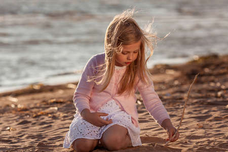 Portrait of little cute girl drawing with a stick on the sand at the sunset beachの写真素材