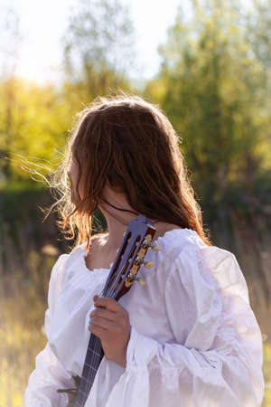 Back view of young woman with red hair in a white medieval dress. Hippie girl holding acoustic guitar at nature.の写真素材