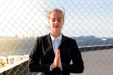 attractive young red-haired business woman in an office suit stands in a yoga pose of relaxation on a background of a metal grid and a sunny cityscape behind herの写真素材