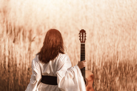 Beautiful young woman with red hair in a white medieval dress holding guitar and walking through the...