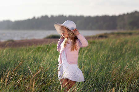Cute little girl in white dress and hat looking to the sea among green field of grass in sunsetの写真素材
