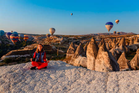 CAPPADOCIA, TURKEY â AUGUST 23, 2019: Young beautiful sitting girl photographer with camera in her hands and flight of hot air balloons at sunrise in Cappadocia. Goreme, Cappadocia, Turkeyのeditorial素材