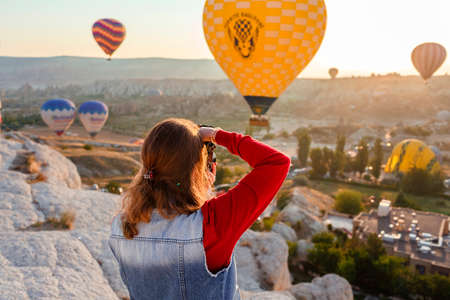 CAPPADOCIA, TURKEY â AUGUST 23, 2019: Young beautiful girl photographer with camera in her hands and flight of hot air balloons at sunrise in Cappadocia. Goreme, Cappadocia, Turkeyのeditorial素材