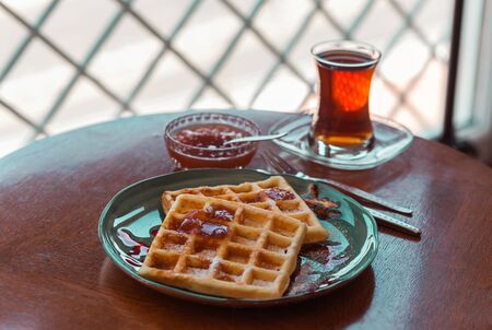 Fresh waffles in sugar powder on a beautiful dish arranged on the table with tea, apricot jam,fork and knife, and decorative lampの写真素材