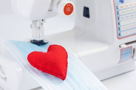 Red heart on mask face fabric. Health care and coronavirus protect with sew mask. Sewing medical masks to protect against coronavirus.Production of masks on sewing machine during quarantineの写真素材